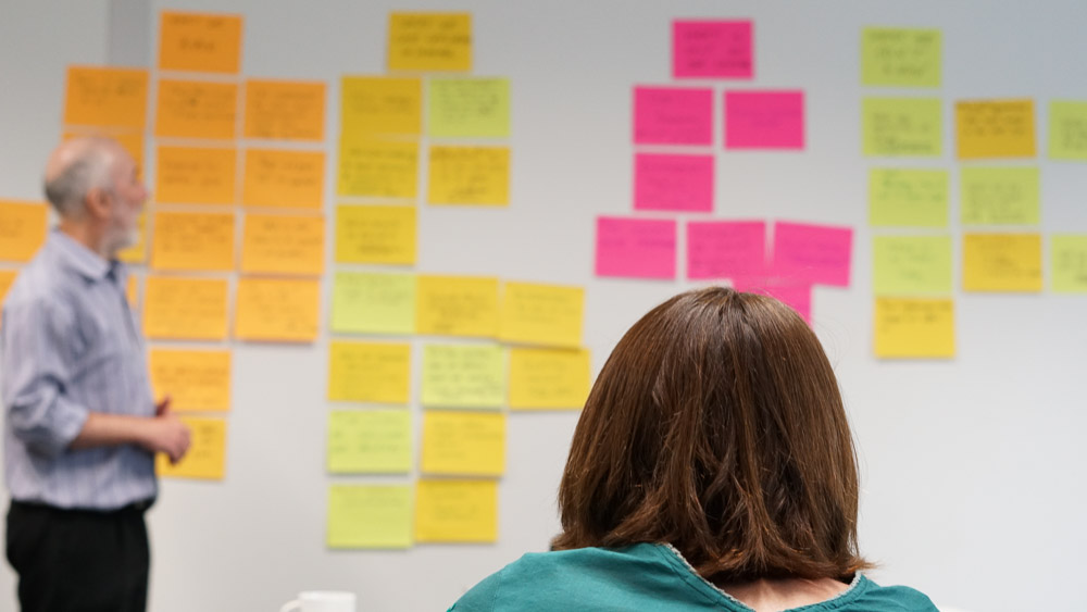Back of a person's head looking at a whiteboard covered in orange, yellow and pink sticky notes while a man stands to the left looking at the board
