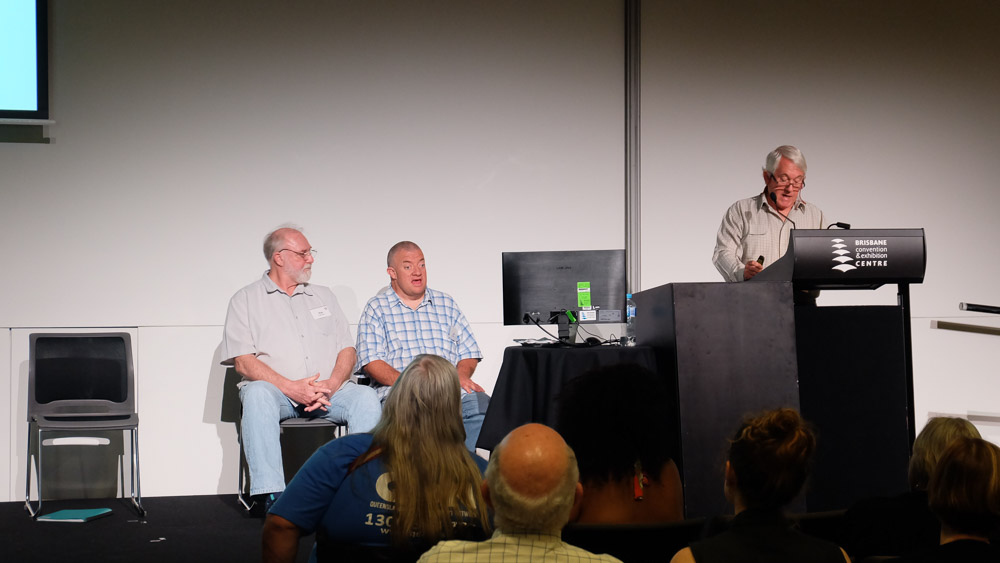 A man speaking at a podium to a room of people, two men are sitting being the speaker