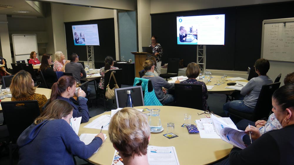 People sitting around round tables watching a powerpoint presentation and listening to a speaker