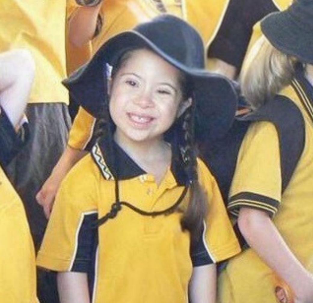 Young girl with braided hair wearing yellow school uniform and wide brimmed hat while grinning.